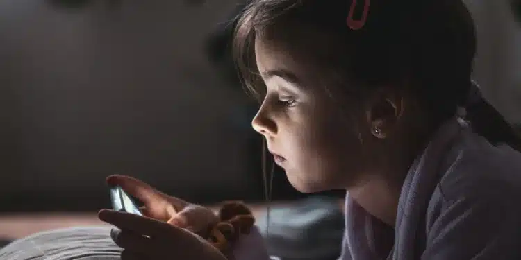 "Menina pequena usando um smartphone na cama, focada na tela, representando a questão do uso de dispositivos por crianças.