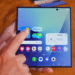 Mão interage com smartphone dobrável exibindo tela inicial e widgets