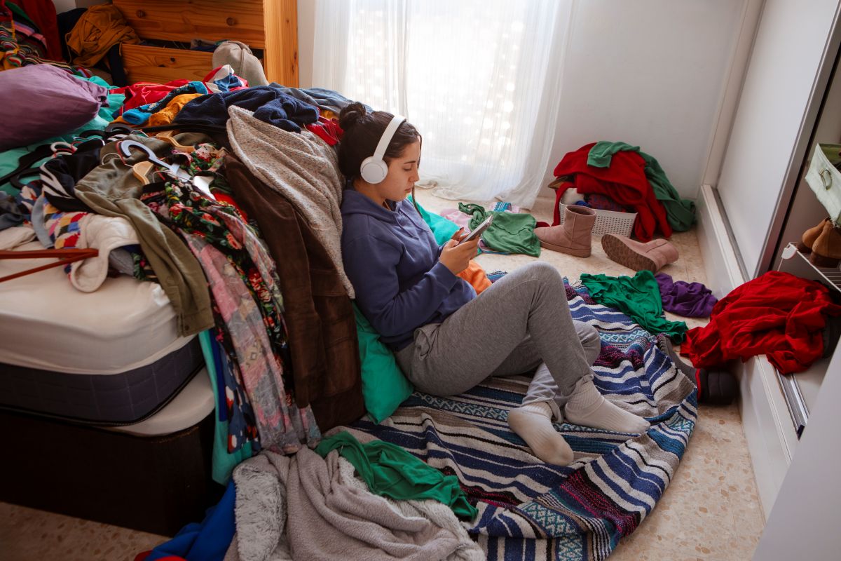 Pessoa sentada no chão de um quarto cercada por roupas espalhadas enquanto usa celular, ilustrando a relação entre bagunça doméstica e saúde emocional.