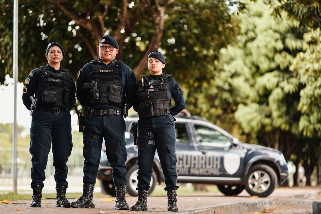 Três guardas municipais uniformizados posam ao lado de viatura, cercados por árvores