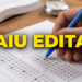 Mão preenchendo gabarito de prova ao lado de livro aberto e cronômetro na mesa