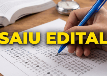 Mão marca alternativa em gabarito de prova objetiva com caneta azul e livro aberto ao fundo
