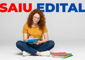 Jovem sentada escrevendo em caderno com edital aberto ao fundo e materiais escolares.