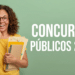 Mulher segurando cadernos e folders amarelos sorri diante de parede verde com texto Concursos Públicos 2026