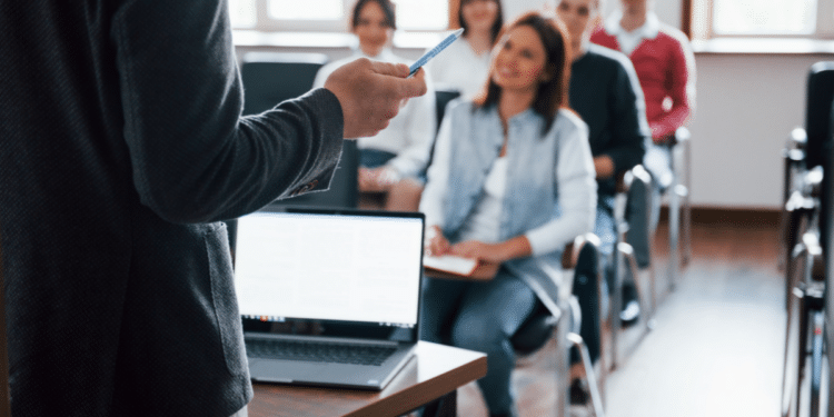 Homem em pé apresenta conteúdo com caneta em mãos para adultos atentos em sala iluminada