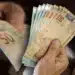 Notas de 50 reais em mãos representando o pagamento do 13º salário do INSS aos beneficiários.