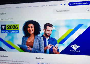 Tela de computador exibindo o portal da Receita Federal com a campanha do Imposto de Renda 2026, com duas pessoas sorrindo ao fundo segurando dispositivos digitais