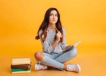 Jovem sentada no chão segura lápis e caderno, olha para cima pensativa ao lado de livros empilhados.