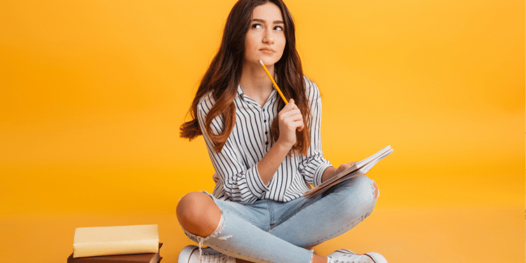 Jovem sentada no chão segura lápis e caderno, olha para cima pensativa ao lado de livros empilhados.