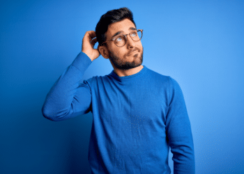 Homem de óculos coçando a cabeça com expressão de dúvida em fundo azul