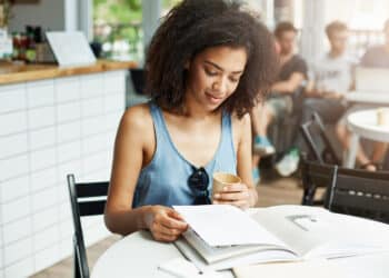 Mulher jovem de cabelo cacheado lendo documento aberto segurando copo em ambiente de cafeteria iluminado