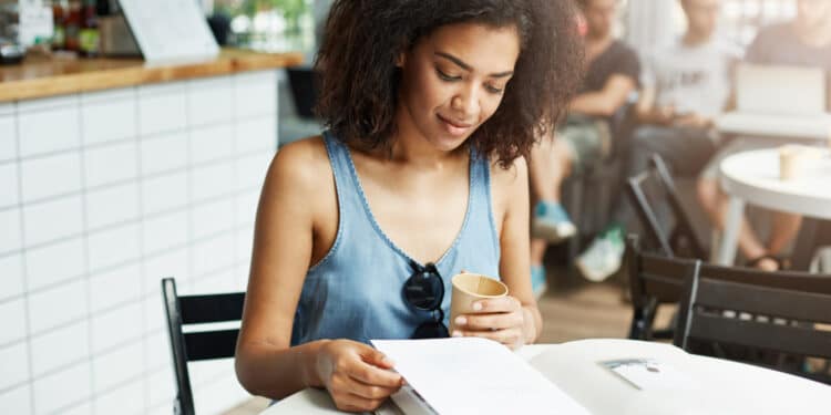 Mulher jovem de cabelo cacheado lendo documento aberto segurando copo em ambiente de cafeteria iluminado