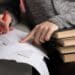 Mãos femininas revisando documentos impressos com lápis vermelho apoiados sobre pilha de livros em ambiente de estudo