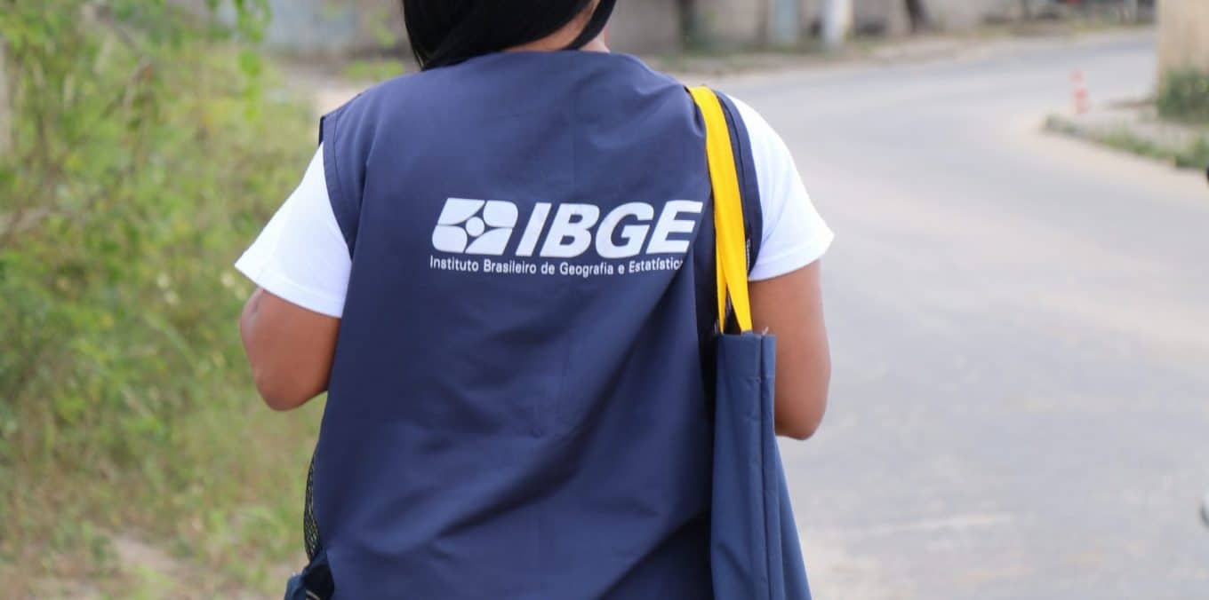 Pessoa com colete do IBGE caminha por rua, carregando bolsa lateral azul