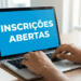 Pessoa digitando em notebook que exibe aviso de inscrições abertas na tela azul