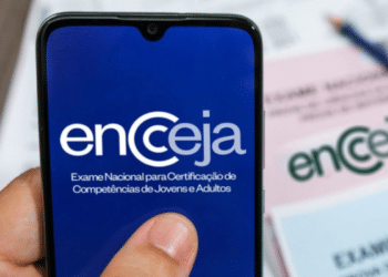 Mão segura celular mostrando logo do Encceja com papéis e caneta ao fundo