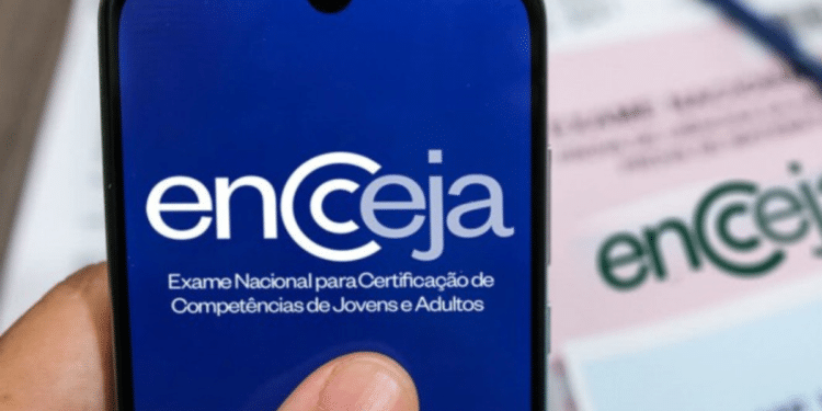 Mão segura celular mostrando logo do Encceja com papéis e caneta ao fundo