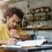 Homem jovem com cabelo afro, usando camiseta amarela, concentrado fazendo anotações em um livro com um notebook ao lado em um ambiente de café.