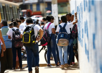 Grupo de estudantes com mochilas reunidos na calçada em frente ao muro escolar.