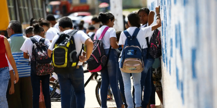 Grupo de estudantes com mochilas reunidos na calçada em frente ao muro escolar.