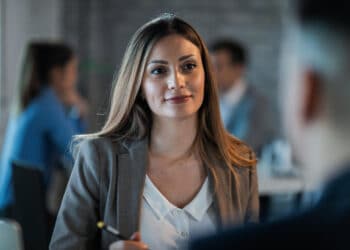 Mulher profissional com blazer cinza e expressão confiante em reunião de trabalho com colegas desfocados ao fundo