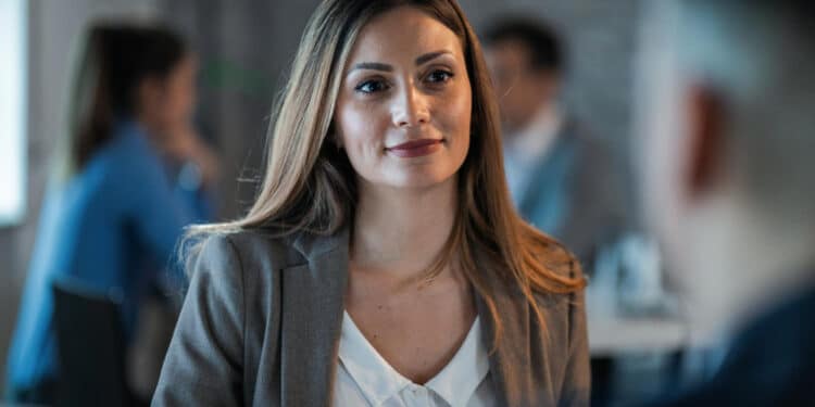 Mulher profissional com blazer cinza e expressão confiante em reunião de trabalho com colegas desfocados ao fundo