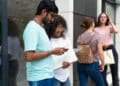 Dois jovens universitários consultando smartphone com notebook em frente a edifício moderno e outras duas estudantes ao fundo em área externa