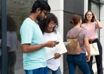 Dois jovens universitários consultando smartphone com notebook em frente a edifício moderno e outras duas estudantes ao fundo em área externa