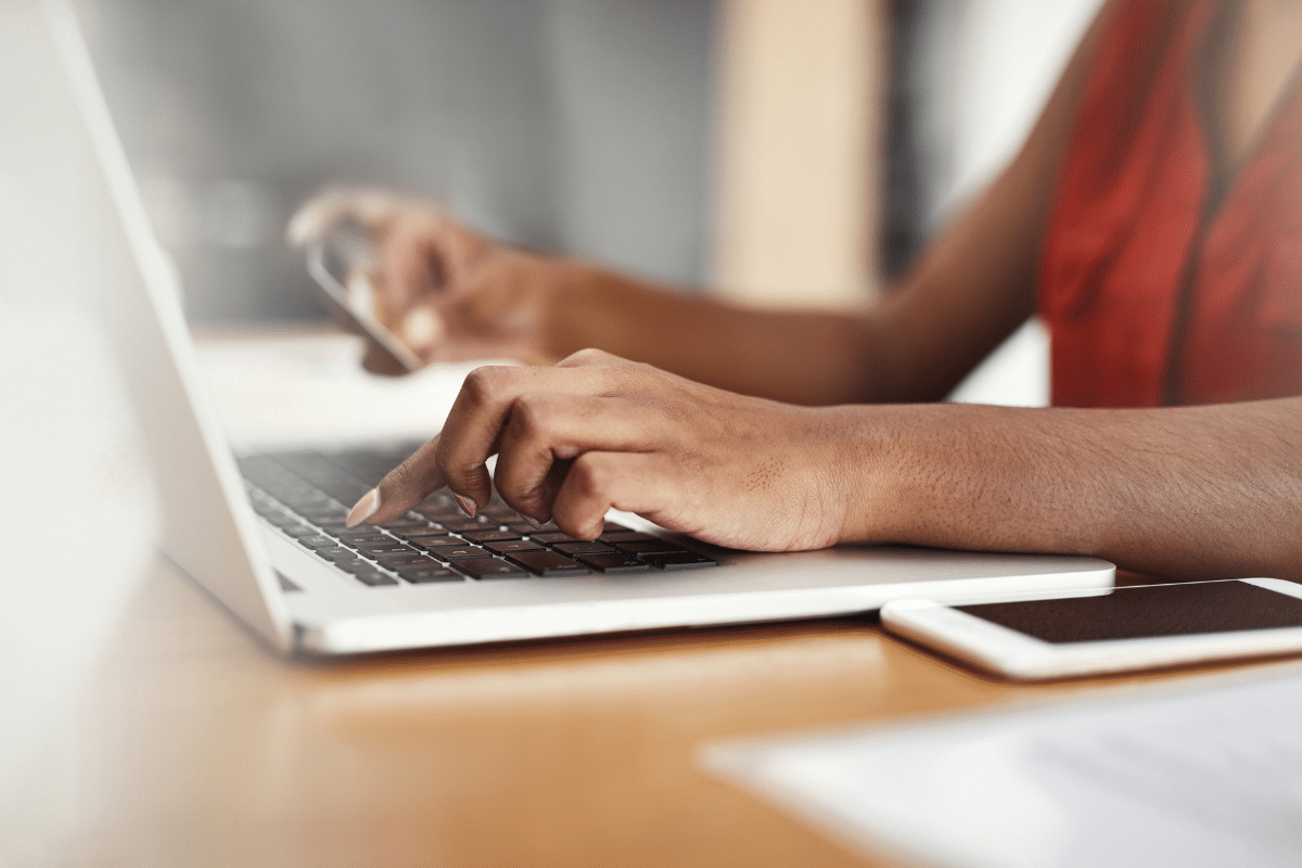 Pessoa utilizando notebook enquanto segura celular, com tablet ao lado na mesa