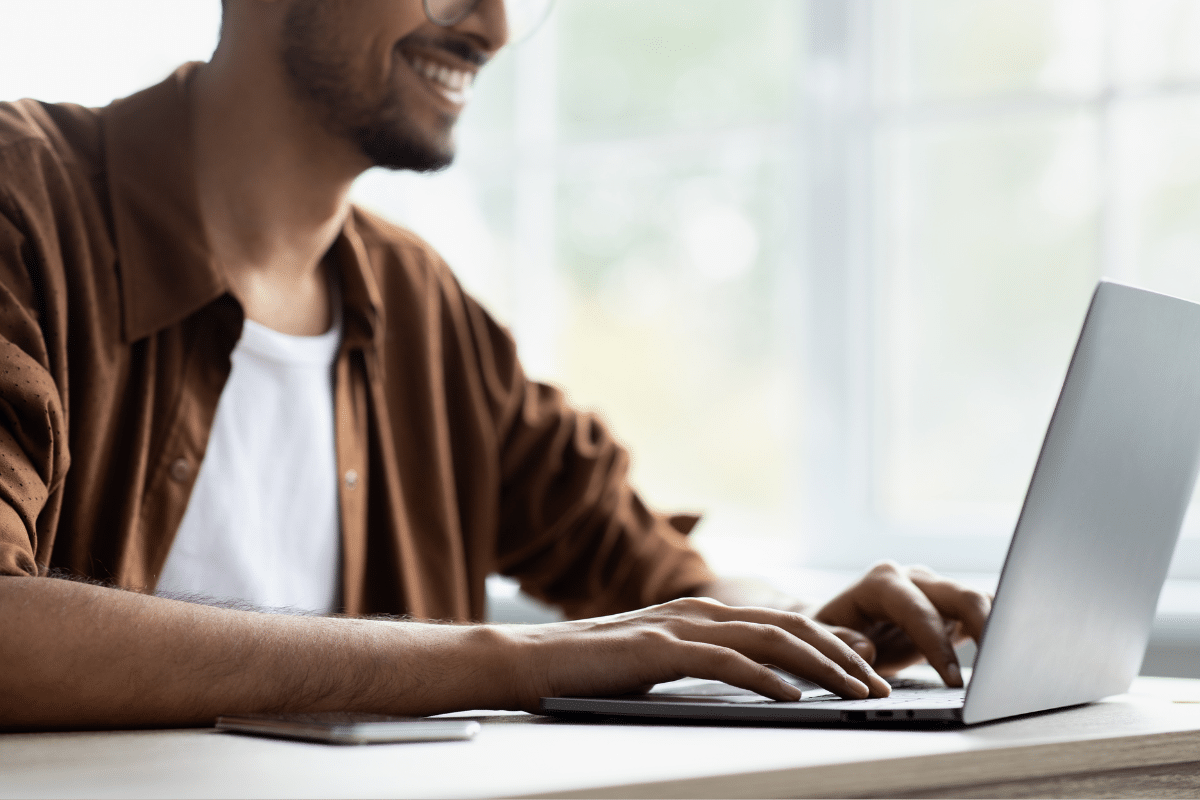 Homem sorridente usando notebook sobre mesa próxima à janela iluminada