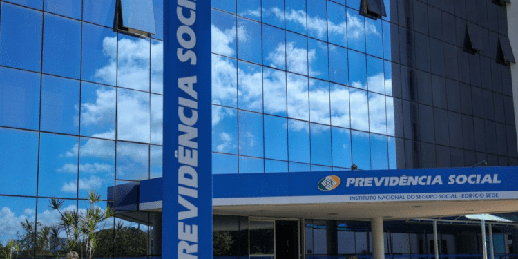 Entrada do prédio do INSS com letreiro azul da Previdência Social e reflexo de nuvens no vidro