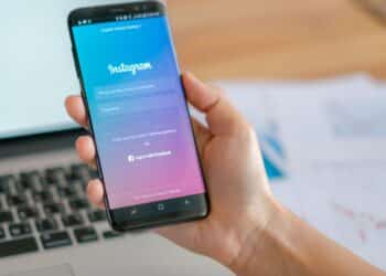 Mão segurando um smartphone com a tela de login do Instagram aberta sobre uma mesa de escritório com um notebook.