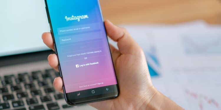 Mão segurando um smartphone com a tela de login do Instagram aberta sobre uma mesa de escritório com um notebook.