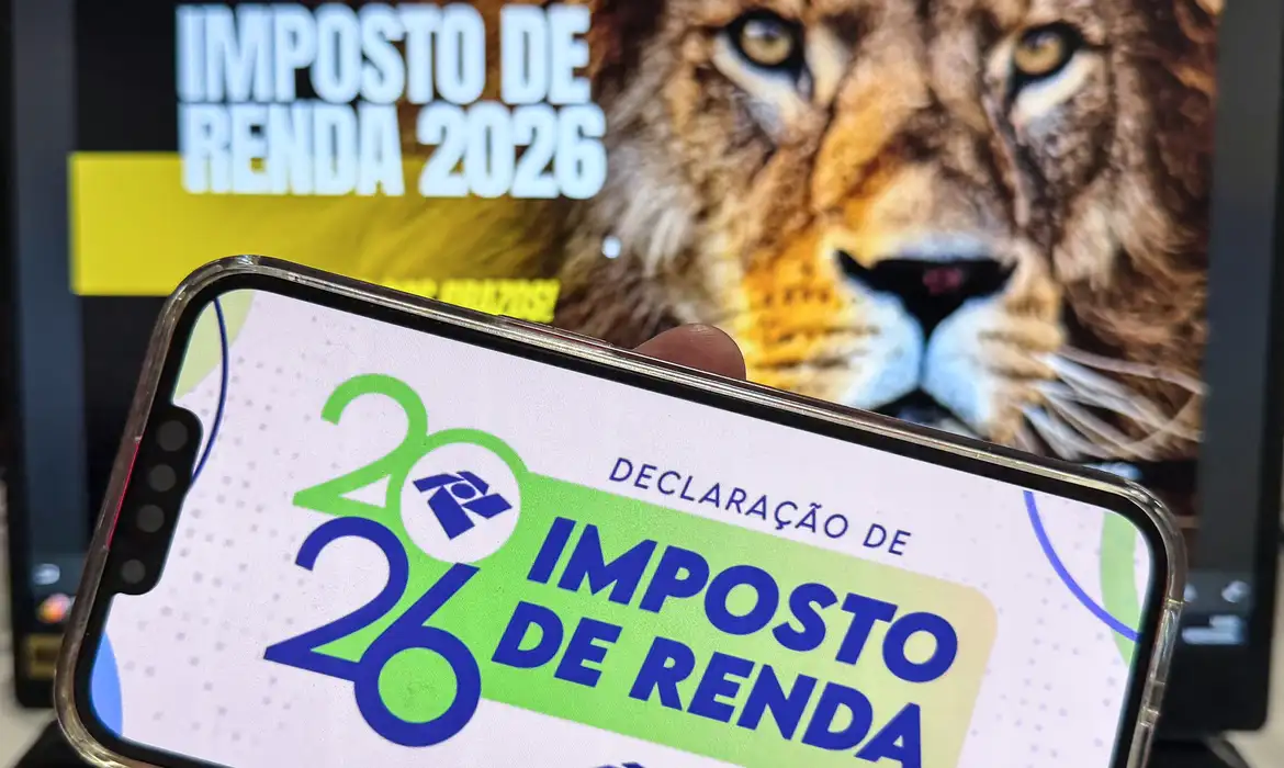 Celular exibindo a declaração do Imposto de Renda 2026 com o leão da Receita Federal ao fundo, simbolizando a fiscalização das declarações.