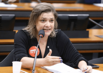 Mulher fala ao microfone durante reunião, gesticulando com a mão e usando crachá sobre projeto