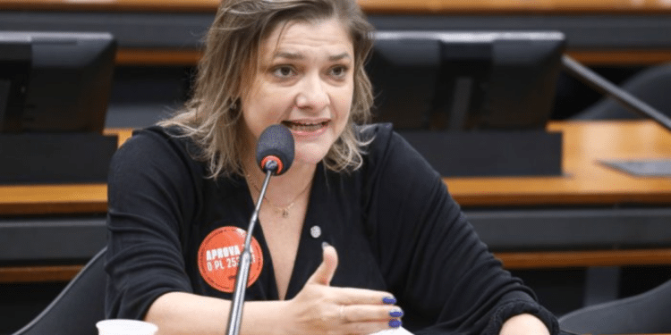 Mulher fala ao microfone durante reunião, gesticulando com a mão e usando crachá sobre projeto