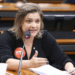 Mulher fala ao microfone durante reunião, gesticulando com a mão e usando crachá sobre projeto