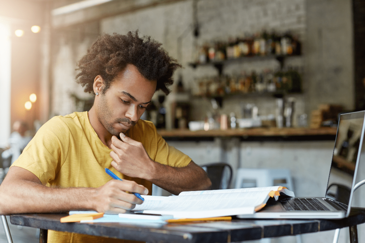 Estudante concentra-se em anotações e livros abertos ao lado de notebook em mesa de café