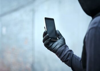 Pessoa com o rosto coberto segurando um smartphone, representando o risco de roubo de celulares e a importância de funções de segurança digital.