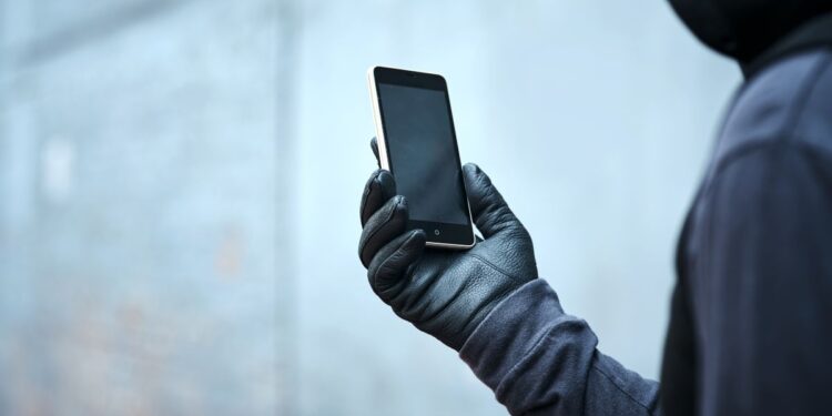 Pessoa com o rosto coberto segurando um smartphone, representando o risco de roubo de celulares e a importância de funções de segurança digital.