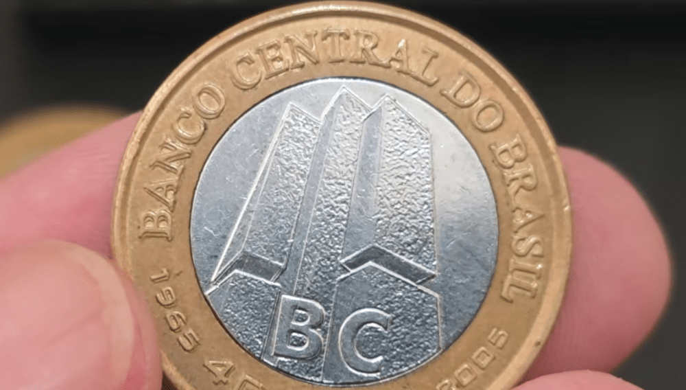 Close-up de uma moeda de 1 Real comemorativa dos 40 anos do Banco Central sendo segurada por dedos. A imagem destaca a matriz de cunhagem com detalhes em relevo no centro prateado e a legenda "BANCO CENTRAL DO BRASIL" no anel dourado, evidenciando possíveis erros de duplicação.