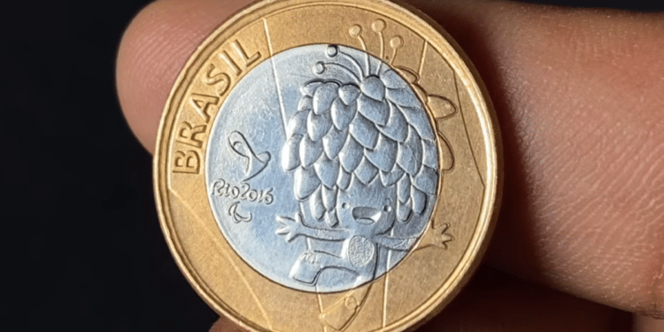 Close-up de uma mão segurando uma moeda de 1 real das Olimpíadas Rio 2016. A face prateada exibe o mascote Tom e a inscrição "Brasil Rio 2016", enquanto a borda é dourada.