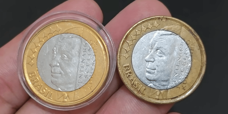 Close de duas moedas de 1 real comemorativas do centenário de Juscelino Kubitschek sendo seguradas entre os dedos. Uma está em estojo protetor e a outra solta.