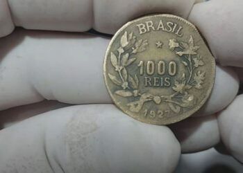 Moeda de bronze-alumínio de 1000 Réis do Brasil, ano 1927, sendo segurada por mãos com luvas brancas de colecionador.