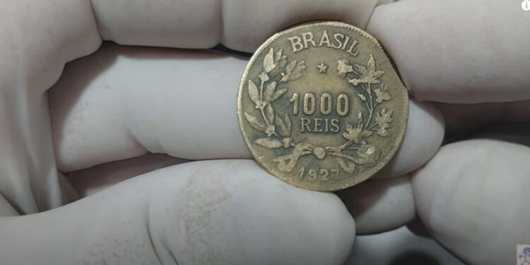 Moeda de bronze-alumínio de 1000 Réis do Brasil, ano 1927, sendo segurada por mãos com luvas brancas de colecionador.