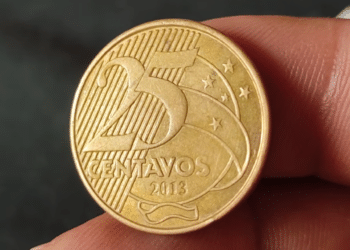 Close em detalhe de uma moeda dourada de 25 centavos de Real, do ano de 2013, sendo segurada pelos dedos de uma pessoa para análise, com foco na data e no valor.