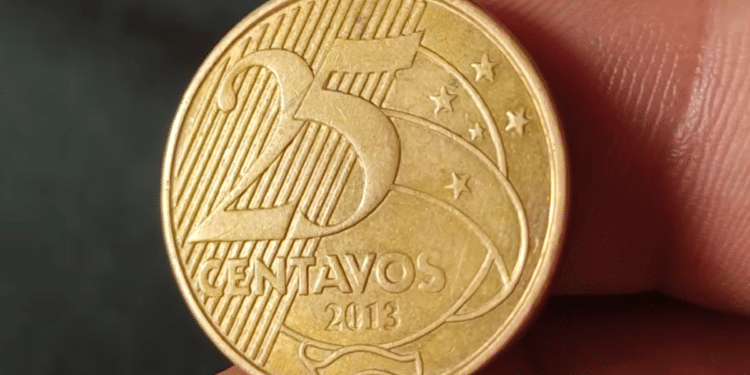 Close em detalhe de uma moeda dourada de 25 centavos de Real, do ano de 2013, sendo segurada pelos dedos de uma pessoa para análise, com foco na data e no valor.