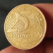 Close em detalhe de uma moeda dourada de 25 centavos de Real, do ano de 2013, sendo segurada pelos dedos de uma pessoa para análise, com foco na data e no valor.