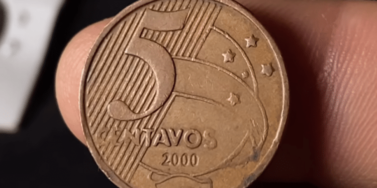 Close do reverso de uma moeda de 5 centavos do ano 2000, de cor bronze, sendo segurada por dedos. O foco está nos detalhes do cunho e na data.