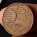 Close do reverso de uma moeda de 5 centavos do ano 2000, de cor bronze, sendo segurada por dedos. O foco está nos detalhes do cunho e na data.